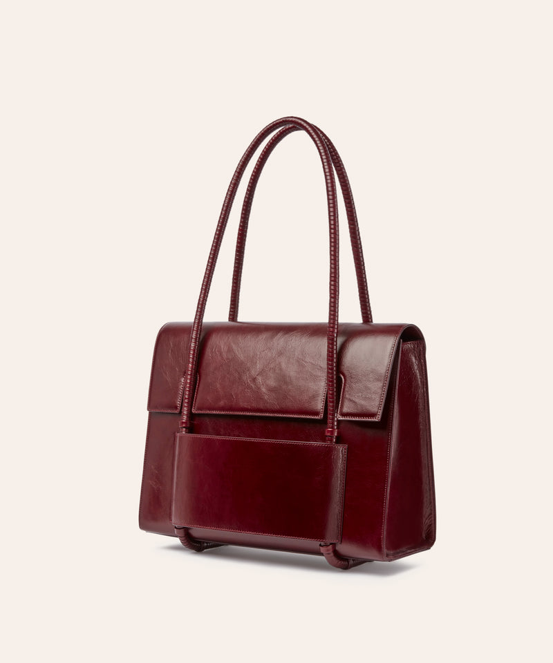 KUN HONG MOTHER SATCHEL LACQUER RED KUN HONG MOTHER SATCHEL Maroon handbag with leather wrapped double handle on a beige background