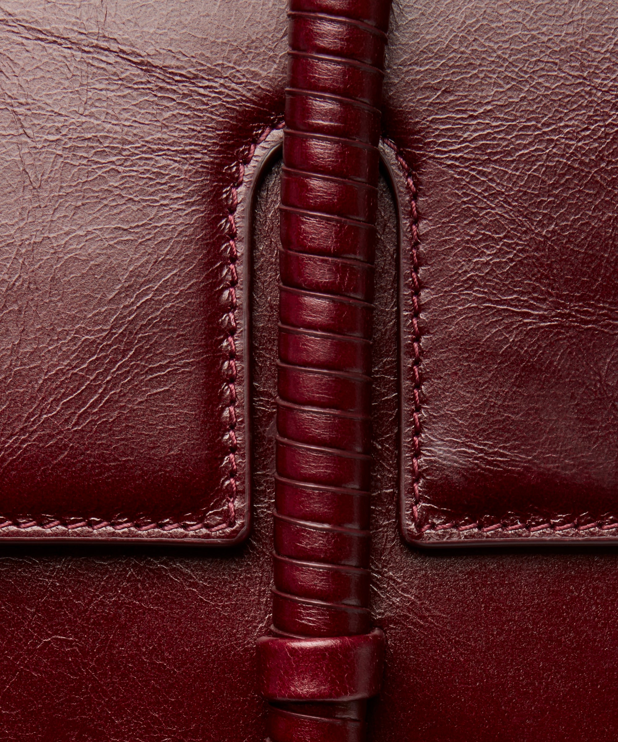 KUN HONG MOTHER SATCHEL LACQUER RED Maroon handbag with leather wrapped double handle on a beige background. Close-up of a brown red burgundy leather bag with leather wrapped handle and stitching detail