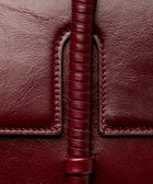 KUN HONG MOTHER SATCHEL LACQUER RED Maroon handbag with leather wrapped double handle on a beige background. Close-up of a brown red burgundy leather bag with leather wrapped handle and stitching detail